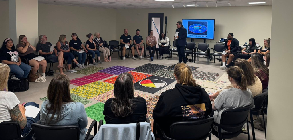 prescott staff sit in a circle with several colourful blankets in the middle while a facilitator speaks to the group. 