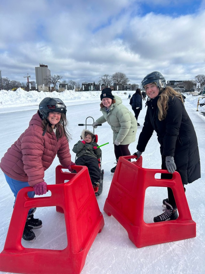 Four diverse people cheerfully ice skate on a bright, sunny rink using specialized frames and adaptive sleds.