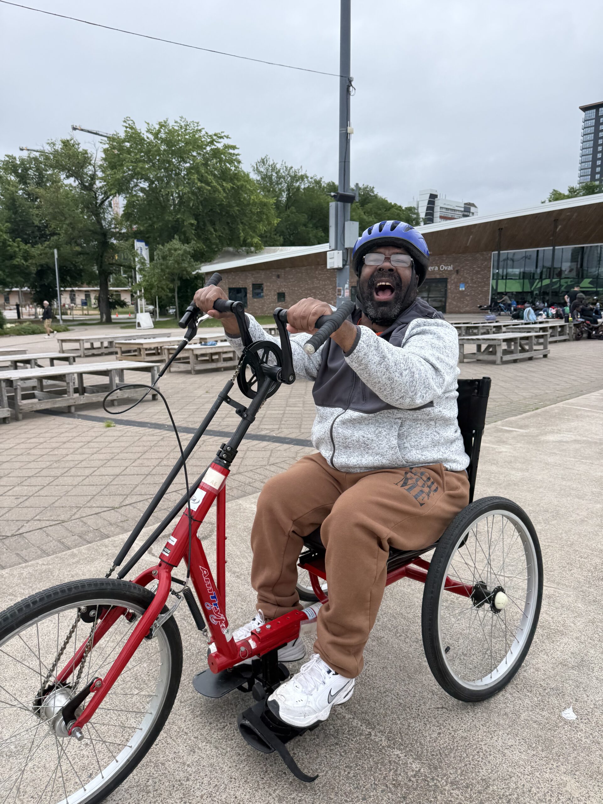 individual smiles while riding a red and black accessible bike with hand petals outside at the emera oval
