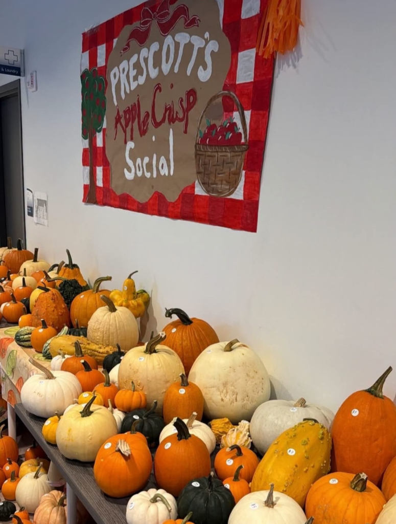small pumpkins and gourds cover a long table with a banner above reading Prescott's apple crisp social